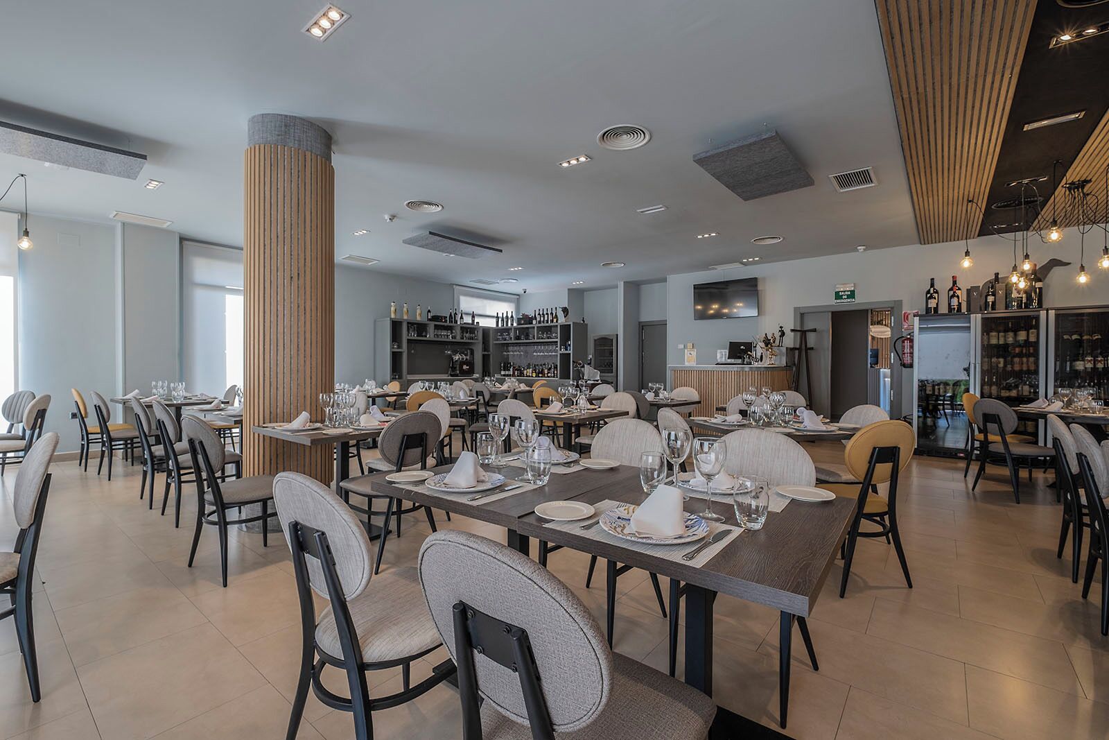 Un comedor amplio con varias mesas dispuestas para comidas, luminarias modernas y un gran pilar en el centro.