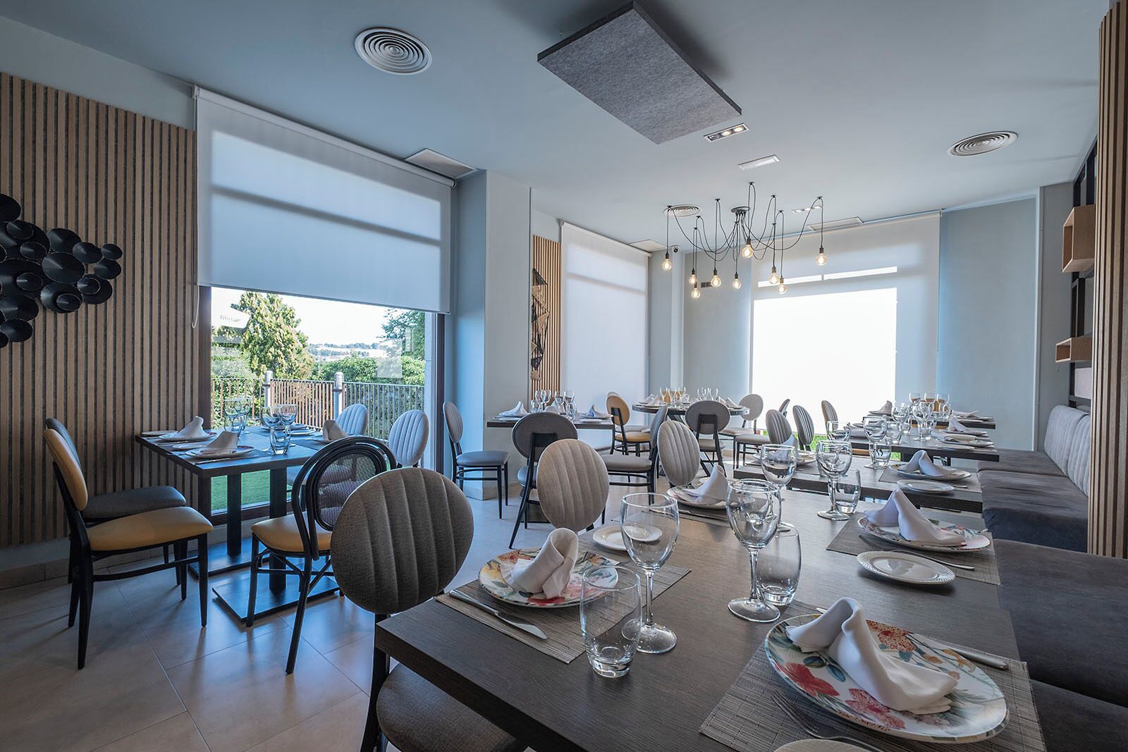 A modern dining area with multiple tables set for guests, large windows offering a view of the outdoors, and contemporary lighting fixtures.