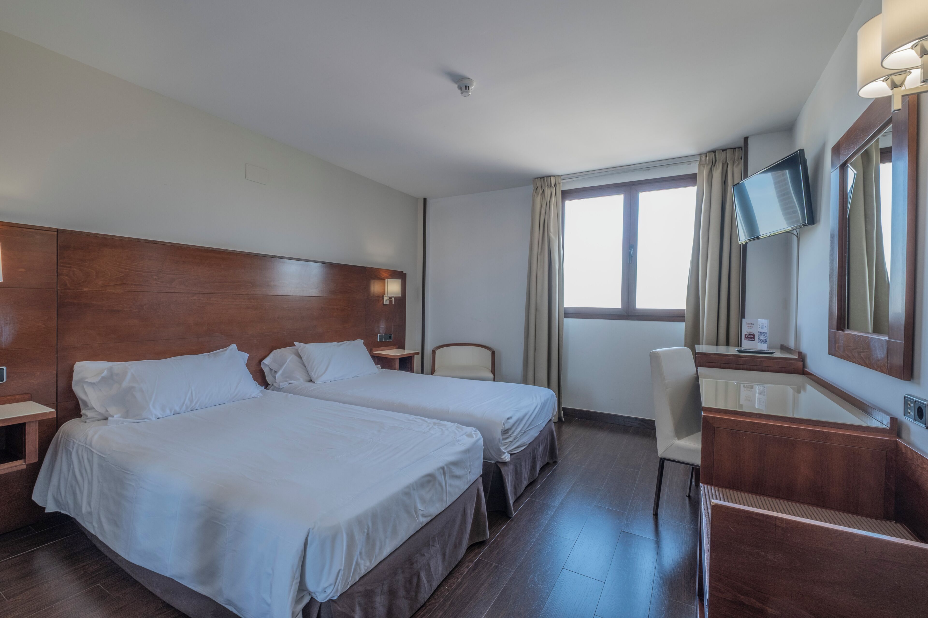 A hotel room with two beds, a desk, a chair, a TV, and a window with curtains.