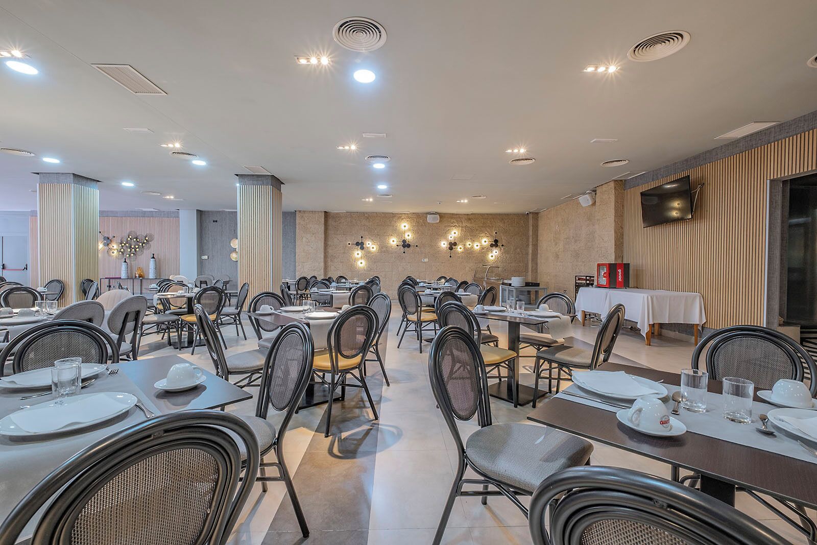 A spacious dining area with multiple tables and chairs, recessed ceiling lights, and a large flat-screen TV mounted on the wall.