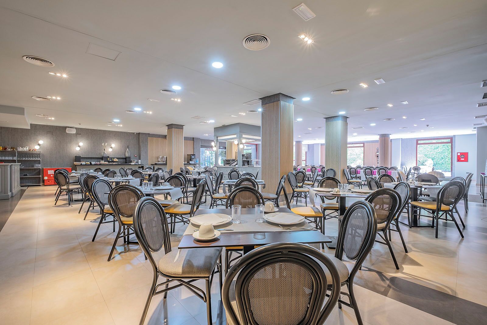 Un grand espace dédié à la salle à manger, avec plusieurs tables et chaises, une cuisine à l’arrière-plan et de grandes fenêtres qui laissent entrer la lumière naturelle.