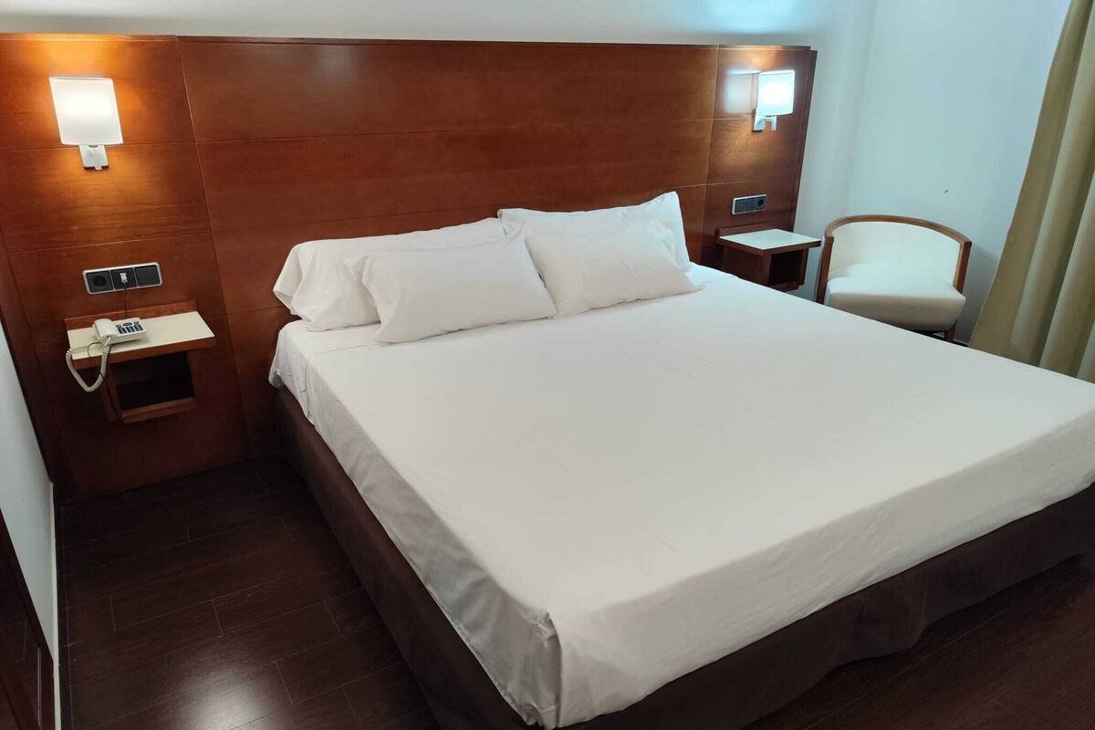 A hotel room with a large bed, wooden headboard, bedside tables with lamps, a telephone, and a chair.