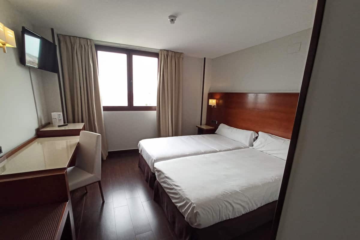 A hotel room with two beds, a desk, a chair, a TV, and a window with curtains.