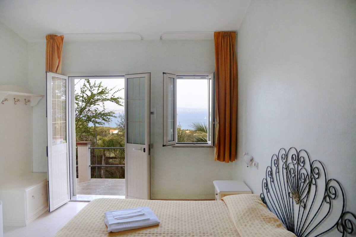 A bedroom with a bed, a window with curtains, a balcony, and a view of the sea.