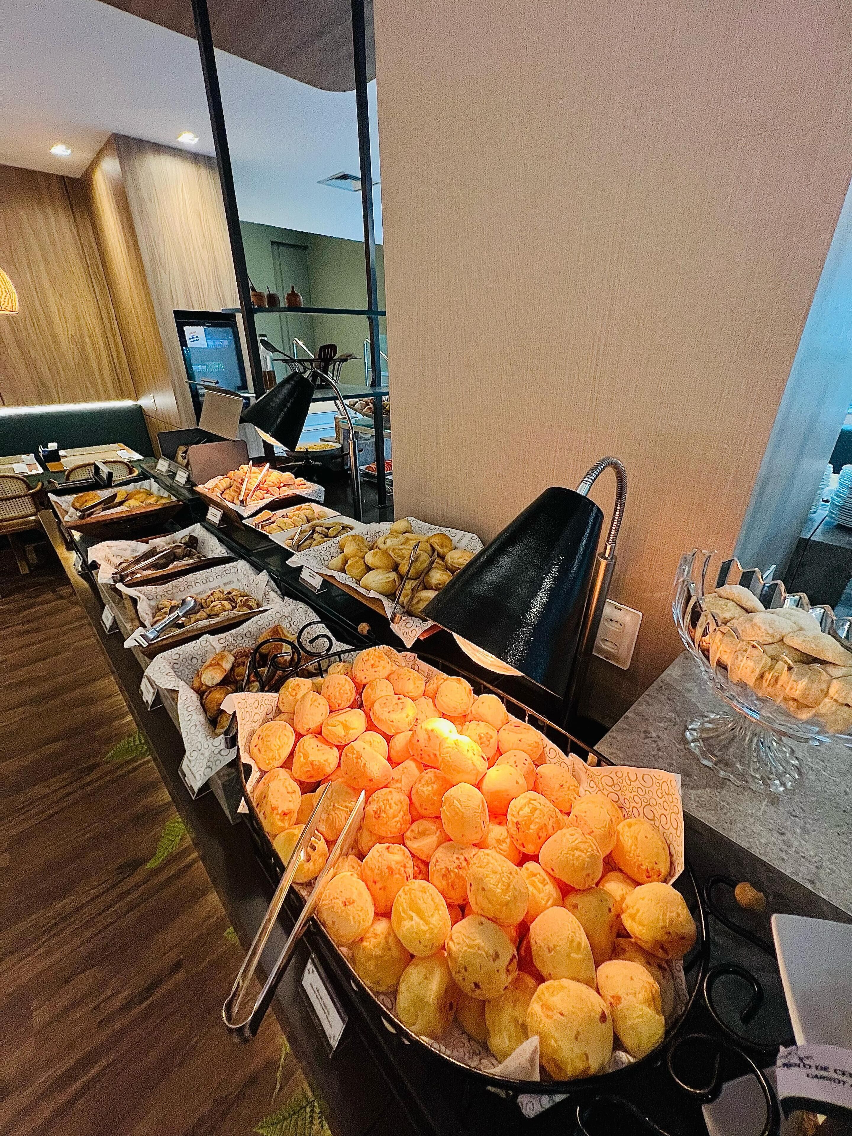 Buffet com diversos alimentos, incluindo pão, doces e uma grande bandeja de itens de cor laranja.