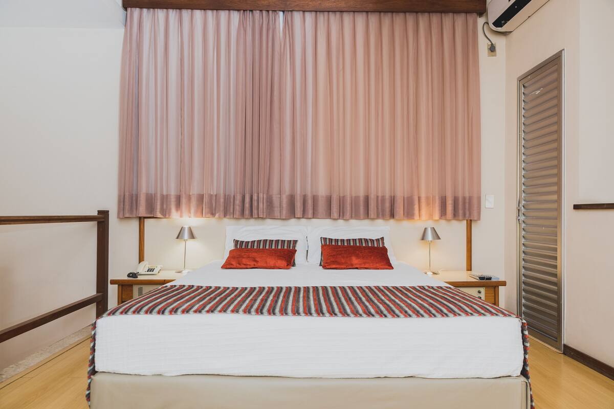 Una cama bien hecha con una colcha a rayas, dos almohadas rojas y un ventanal con cortinas traslúcidas.