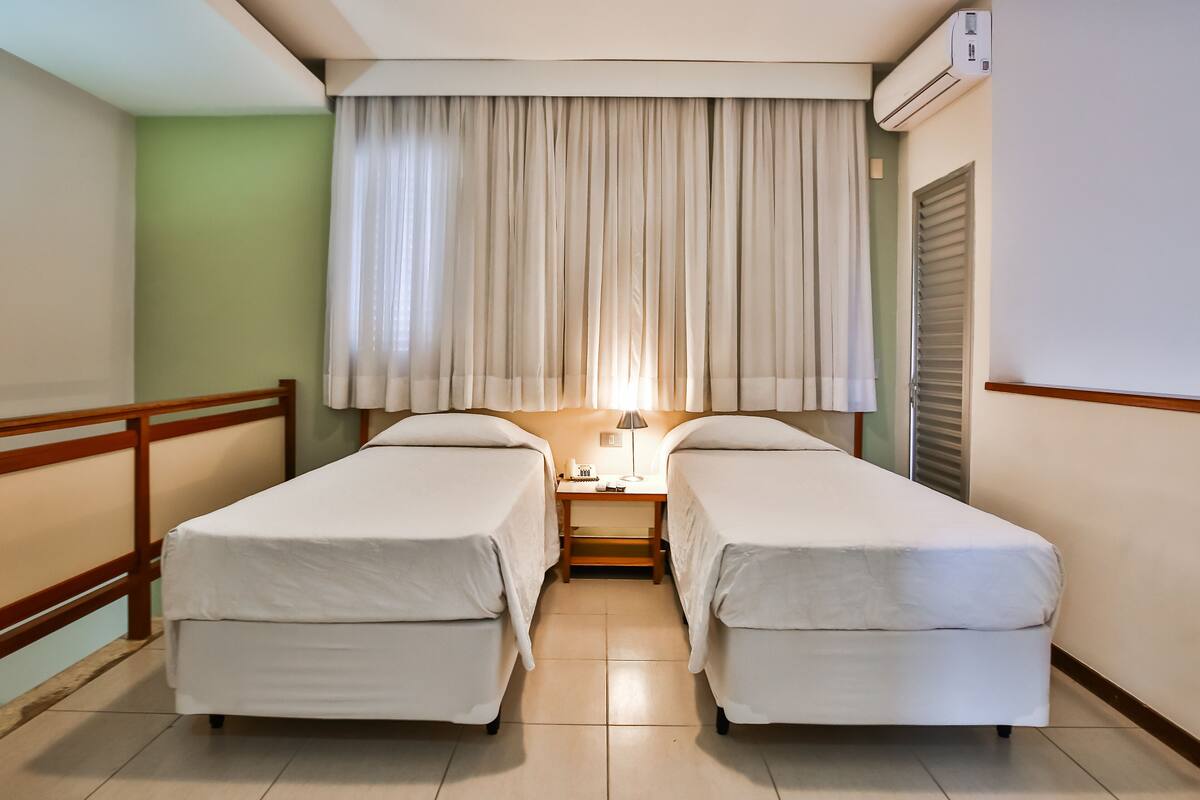 Habitación de hotel con dos camas individuales, una mesita de noche con lámpara, ventana con cortinas y un equipo de aire acondicionado.