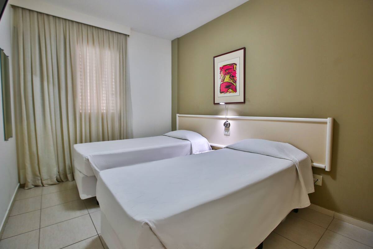 Habitación de hotel con dos camas individuales, un ventanal con cortinas, un cuadro en la pared y una mesita de noche con lámpara.