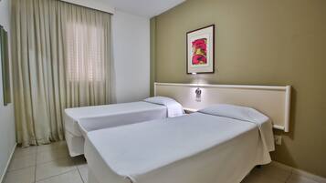 A hotel room with two single beds, a large window with curtains, a framed picture on the wall, and a bedside table with a lamp.