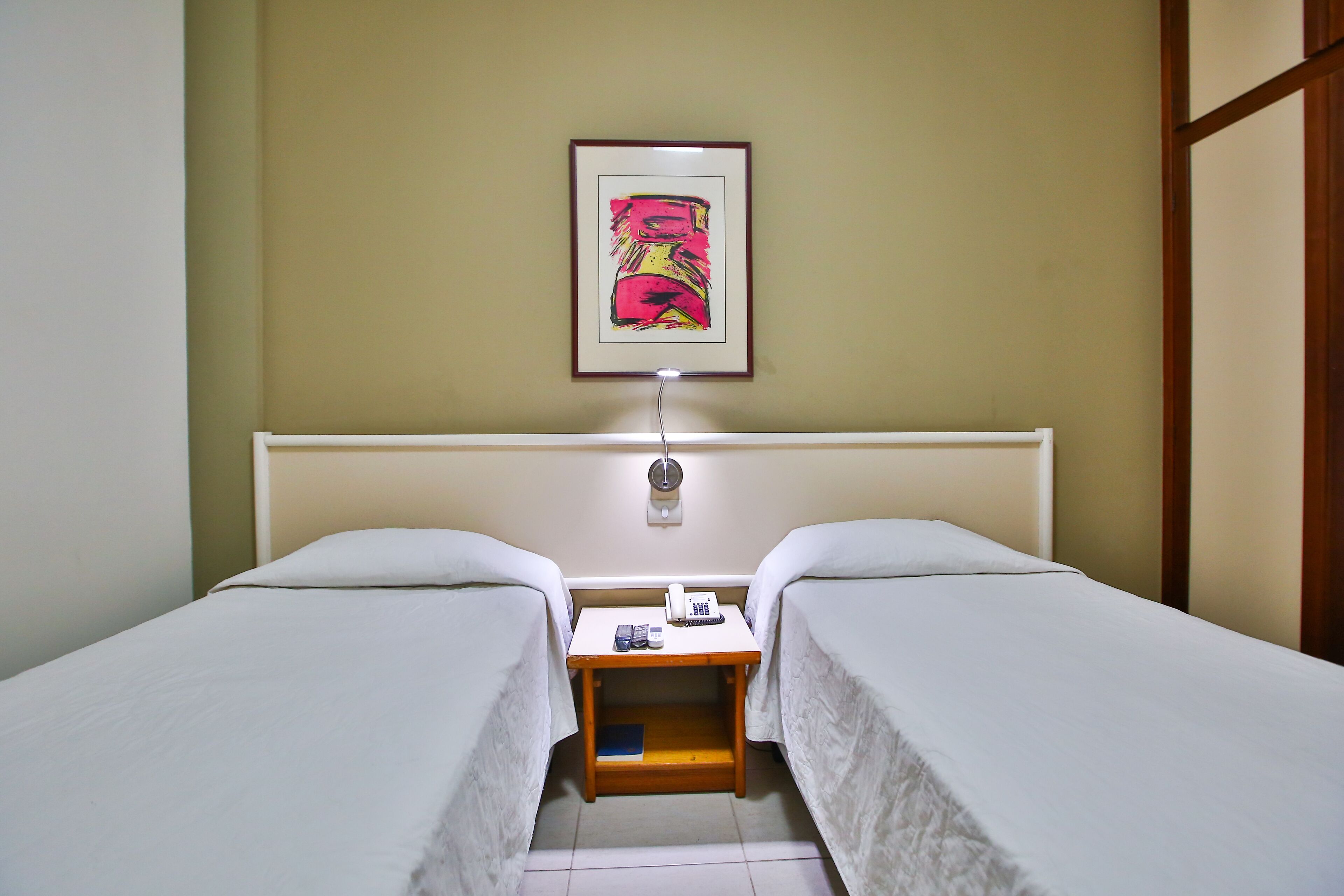 A hotel room with two beds, a bedside table with a lamp, a framed picture on the wall, and a small shelf.