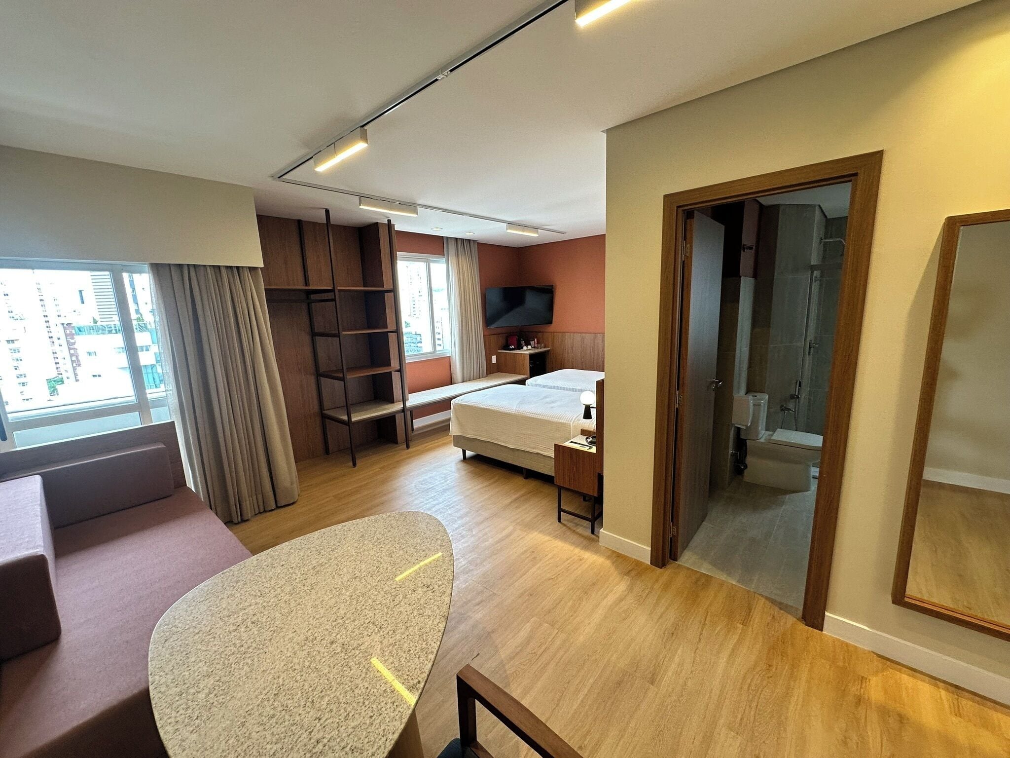 Quarto de hotel moderno com cama, escrivaninha, cadeira, televisão e janela com cortinas.