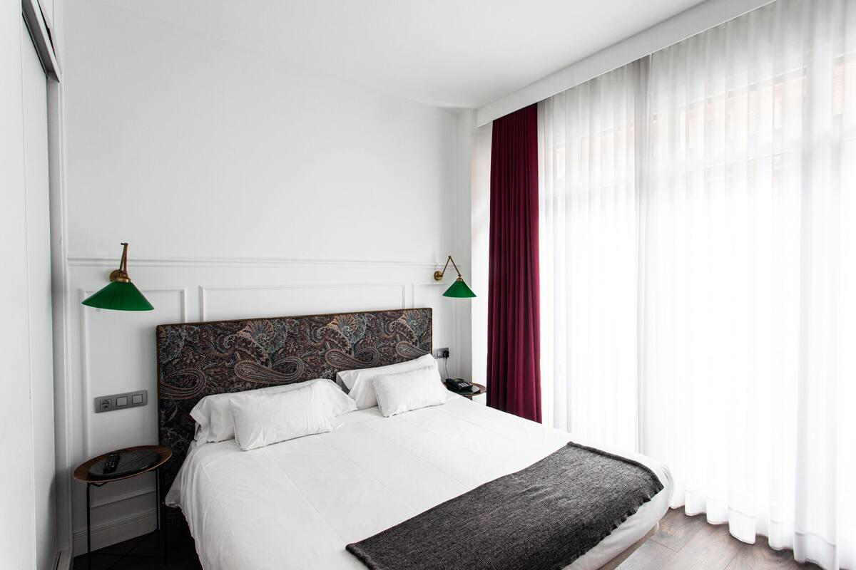 Una cama bien hecha con ropa blanca, un cabecero oscuro con estampado floral, dos lámparas de noche, una mesita de noche con un teléfono y cortinas blancas traslúcidas.