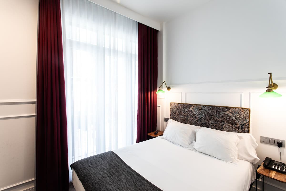 Habitación de hotel con una cama grande, cortinas rojas, un cabecero estampado y dos lámparas de noche.