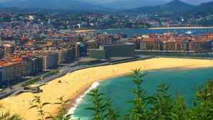 Un paisaje urbano costero con una extensa playa arenosa, un puerto deportivo con barcos y montañas al fondo.