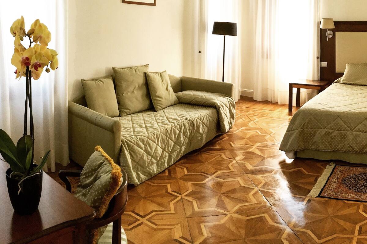Una camera d'albergo con divano, un letto, una scrivania con sedia, una lampada e un quadro appeso al muro.