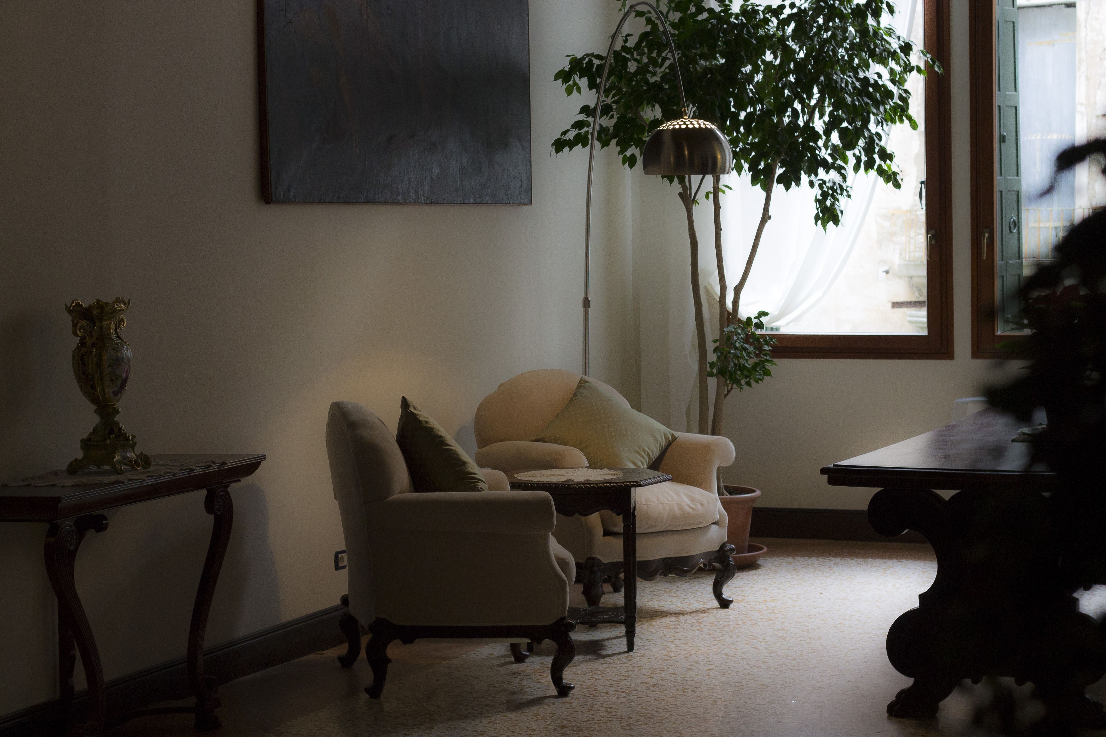 A living room with a beige armchair, a small table, a floor lamp, and a large window with sheer curtains.