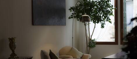 A living room with a beige armchair, a small table, a floor lamp, and a large window with sheer curtains.