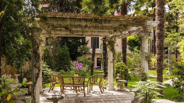A garden patio with a pergola, stone chairs, a table, and a fountain.
