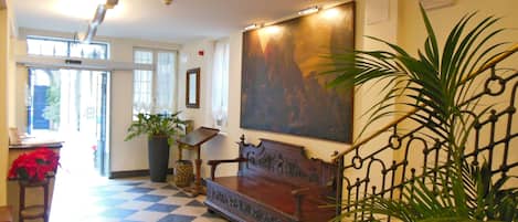 Um hall com chão quadriculado, um banco de madeira, uma pintura grande e um vaso com planta.