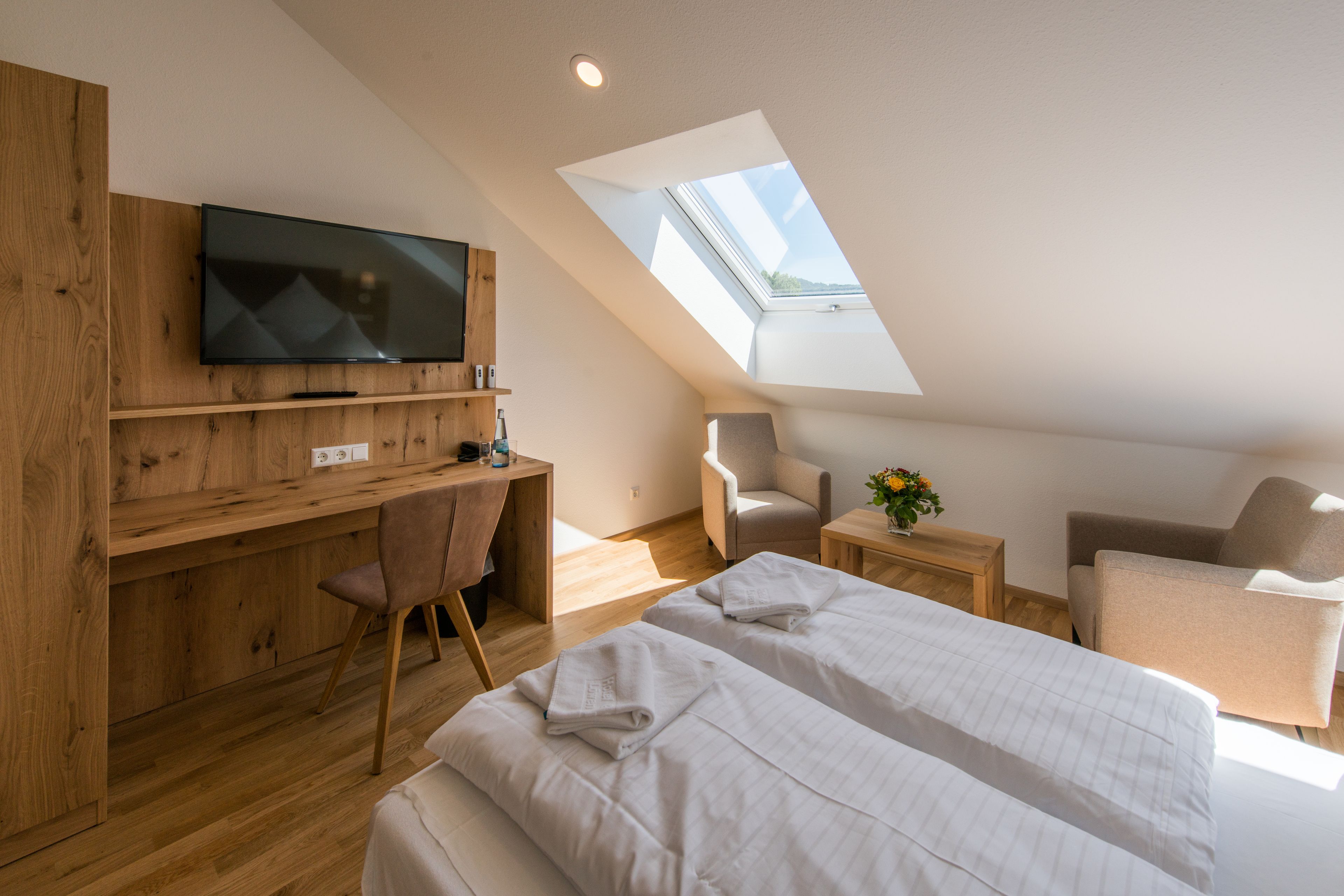 Une chambre moderne avec une lucarne, un téléviseur fixé à un mur en bois, un bureau, deux chaises et un lit avec des serviettes soigneusement pliées.