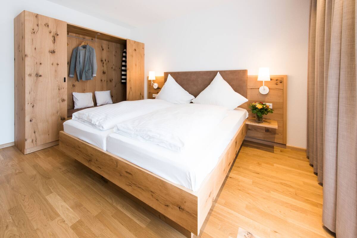 A modern bedroom with a wooden bed frame, a wooden wardrobe, and a bedside table with a lamp and flowers.