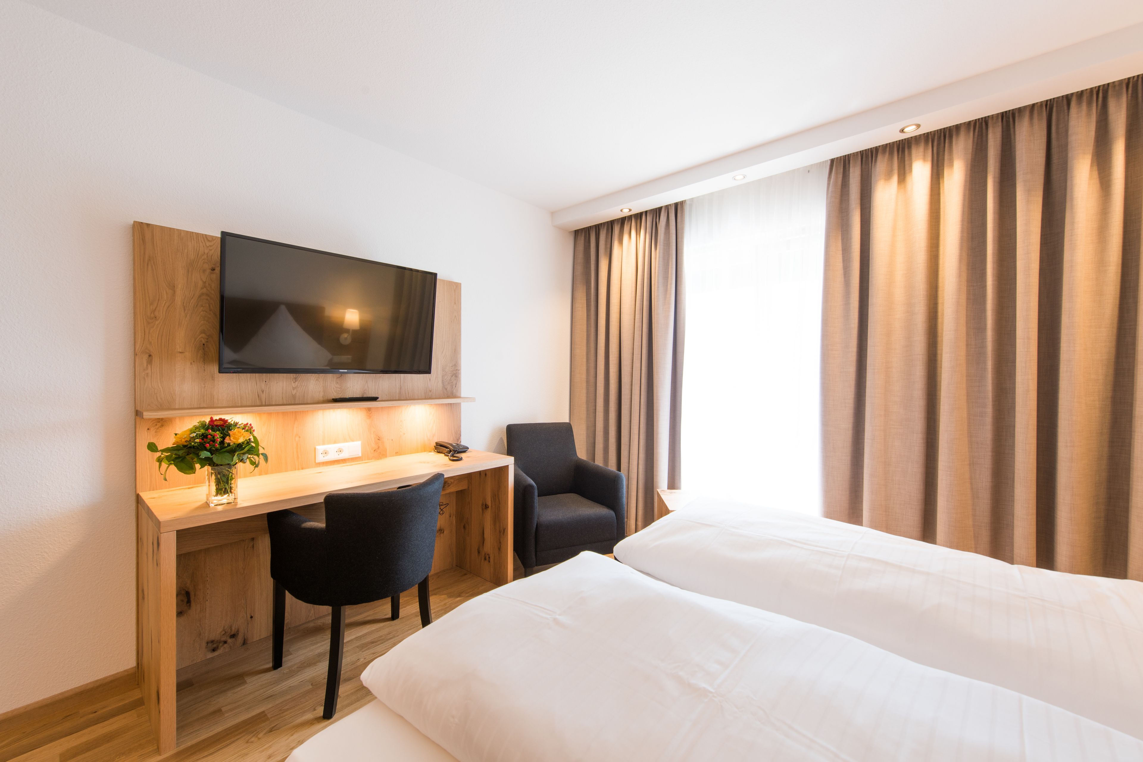 A hotel room with a wooden desk, a flat-screen TV, a black chair, and a bed with white linens.