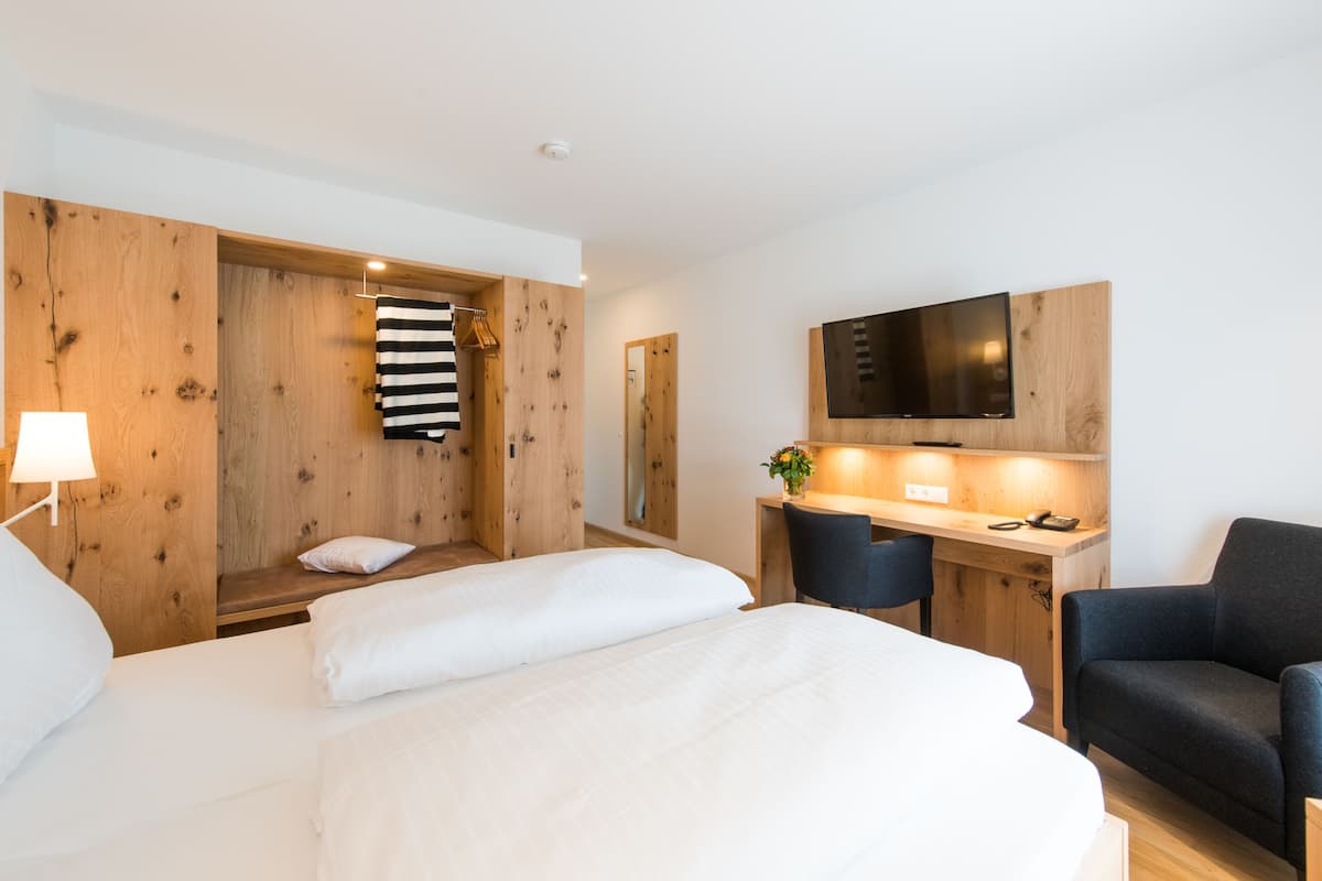 A modern hotel room with a large bed, a desk with a chair, a TV mounted on the wall, and a wooden wardrobe.