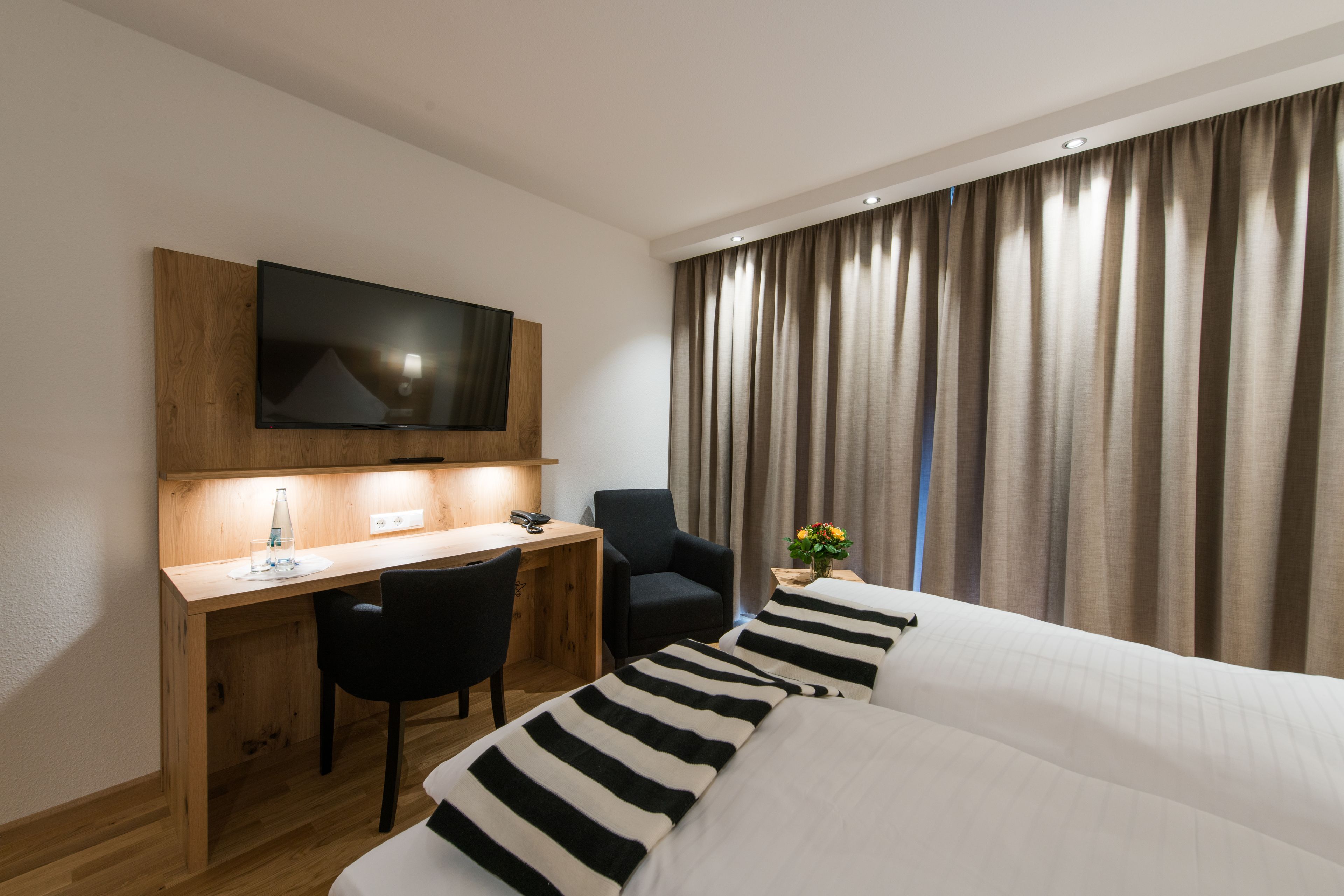 A hotel room with a wooden desk, a black chair, a bed with striped pillows, a TV mounted on the wall, and a window with curtains.