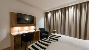 A hotel room with a wooden desk, a black chair, a bed with striped pillows, a TV mounted on the wall, and a window with curtains.