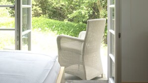 A bedroom with a bed, a wicker chair, and a view of a garden through open windows. - Lieblingsplatz Strandhotel (St. Peter-Ording)