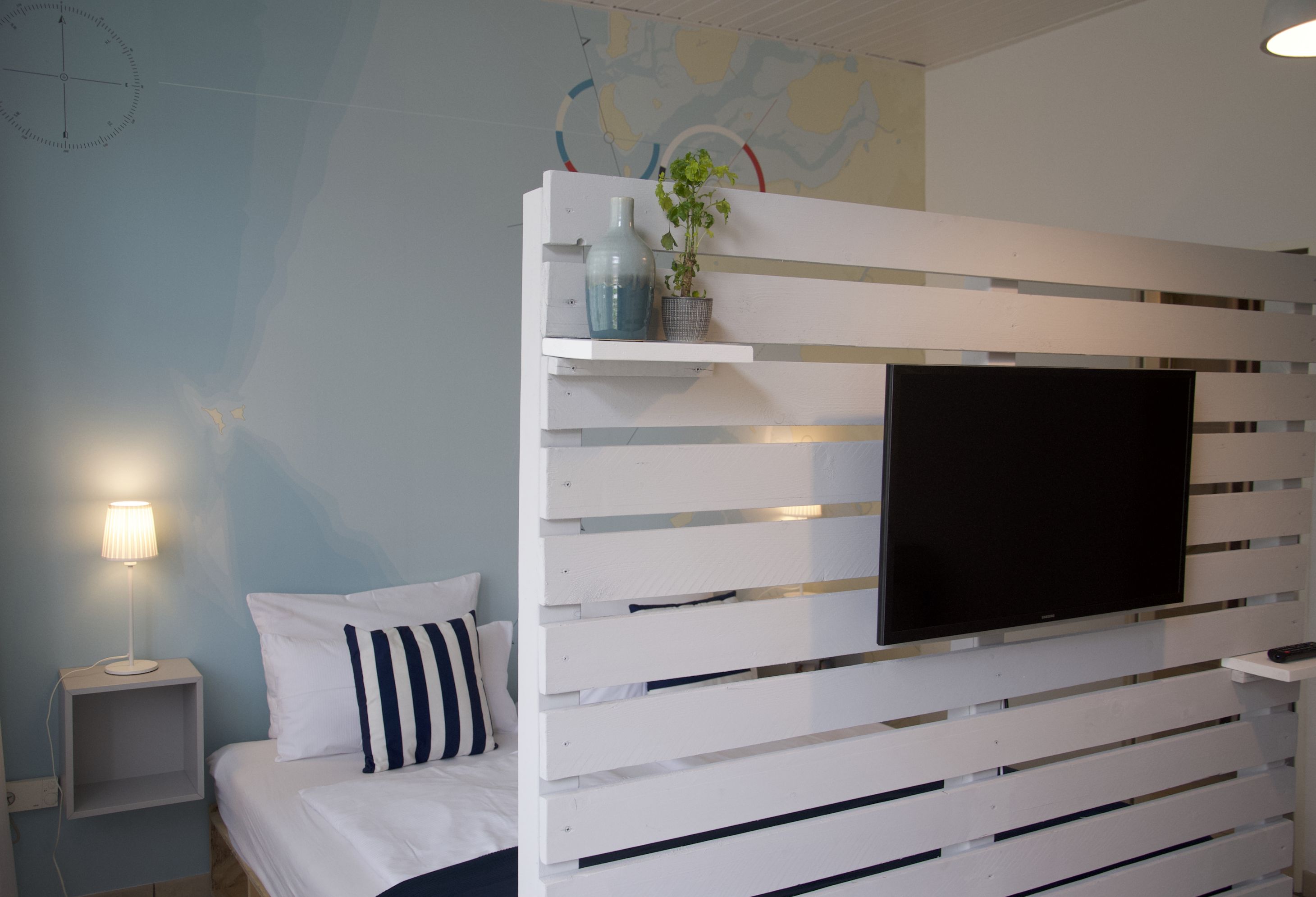 Une chambre à coucher avec un lit, une table de chevet, un téléviseur fixé au mur et une étagère avec une plante.