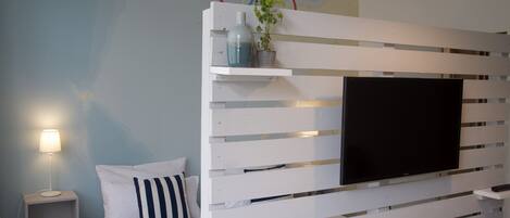 Une chambre à coucher avec un lit, une table de chevet, un téléviseur fixé au mur et une étagère avec une plante.
