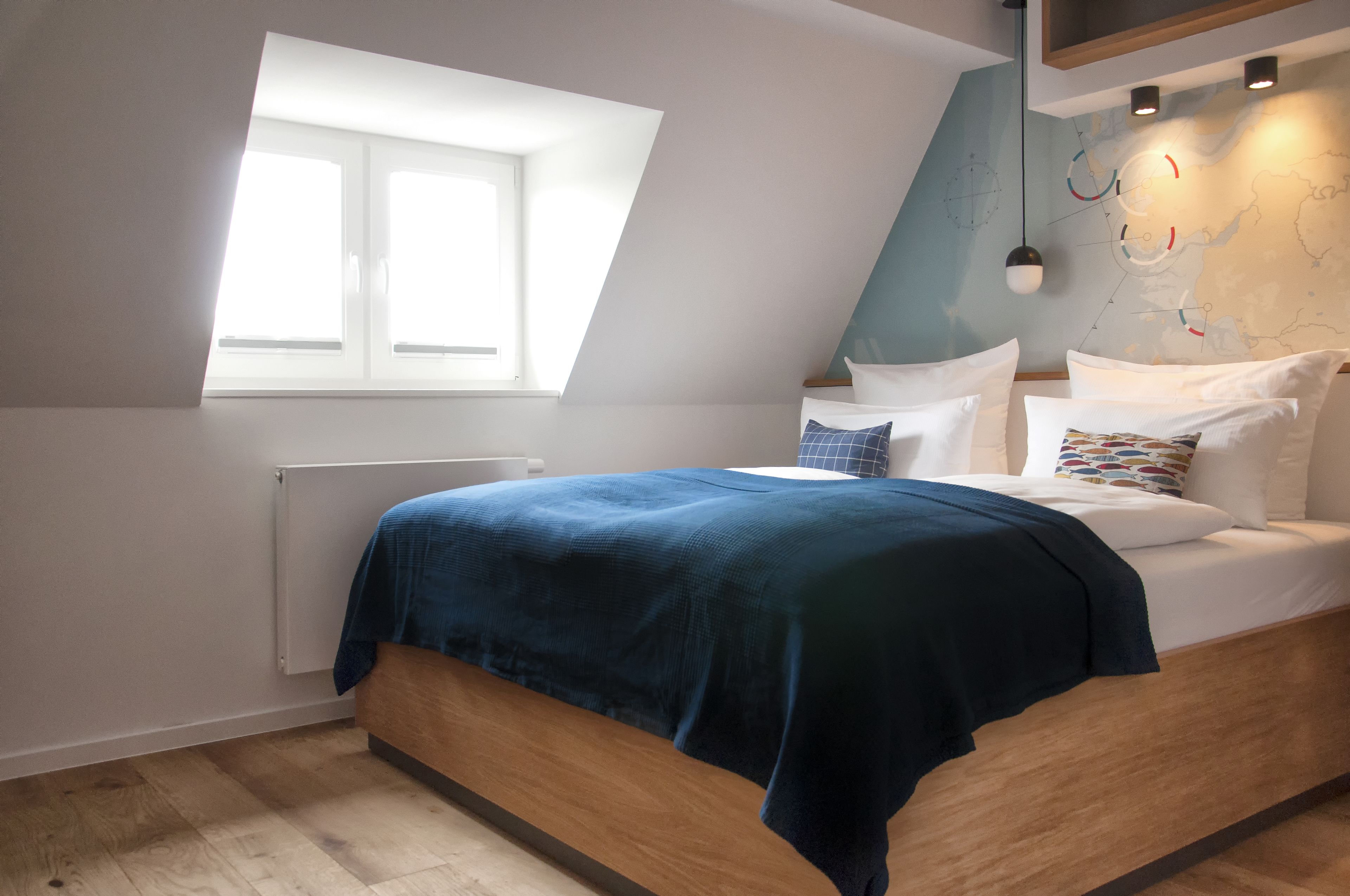 A modern bedroom with a sloped ceiling, a large window, a bed with a blue blanket, white pillows, and a wall map.