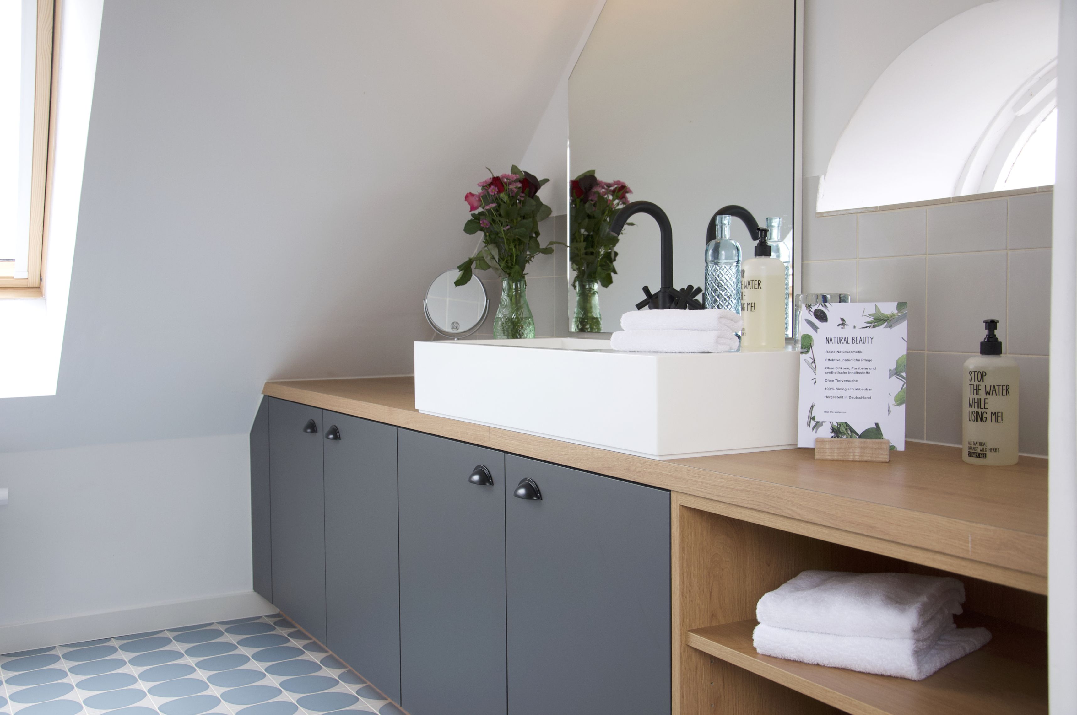 Une salle de bain moderne avec un lavabo blanc, des meubles de couleur gris foncé et un miroir au-dessus du lavabo. On y trouve deux vases avec des fleurs sur le comptoir, ainsi qu’une affiche sur la beauté naturelle.