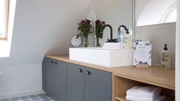 Une salle de bain moderne avec un lavabo blanc, des meubles de couleur gris foncé et un miroir au-dessus du lavabo. On y trouve deux vases avec des fleurs sur le comptoir, ainsi qu’une affiche sur la beauté naturelle.
