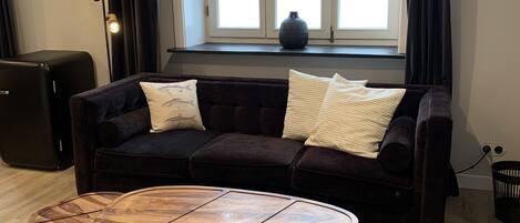 A living room with a dark-colored sofa, a round wooden coffee table, and a large window with curtains.