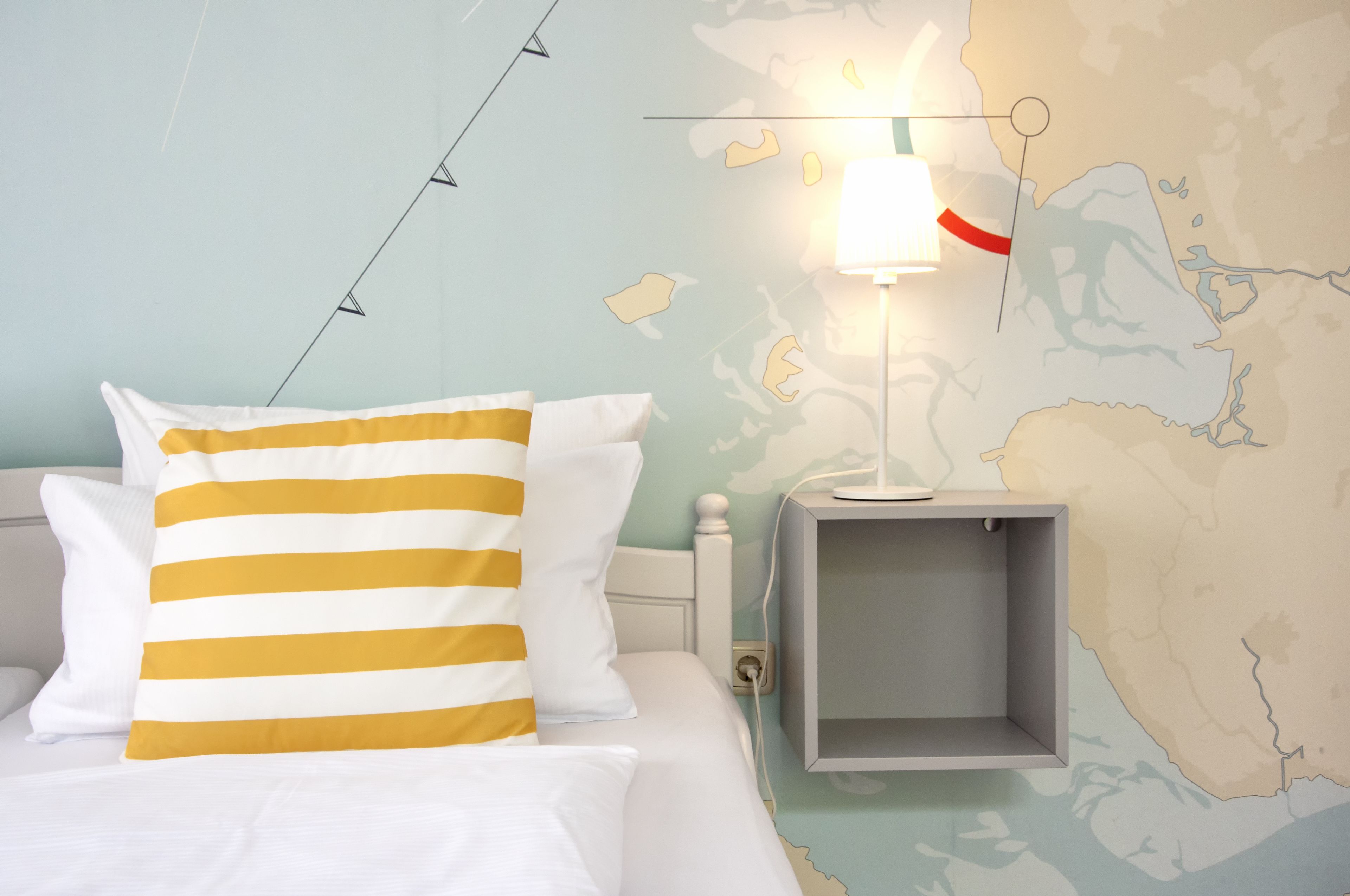A bed with white and yellow striped pillows, a nightstand with a lamp, and a wall map.