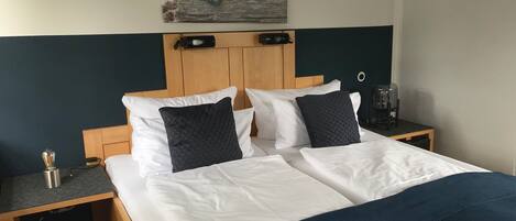 Un lit bien fait, avec des draps blancs et deux oreillers foncés, une tête de lit en bois, une petite table de chevet avec une lampe et un tableau accroché au mur au-dessus du lit.