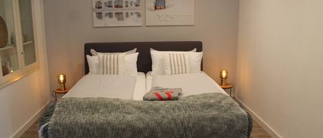 A neatly made bed with white linens, two pillows, and a gray blanket. There are two bedside lamps on either side of the bed.