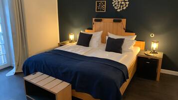 A neatly made bed with a dark blue bedspread, two bedside lamps, a wooden nightstand, a small wooden bench, and a wall art piece.