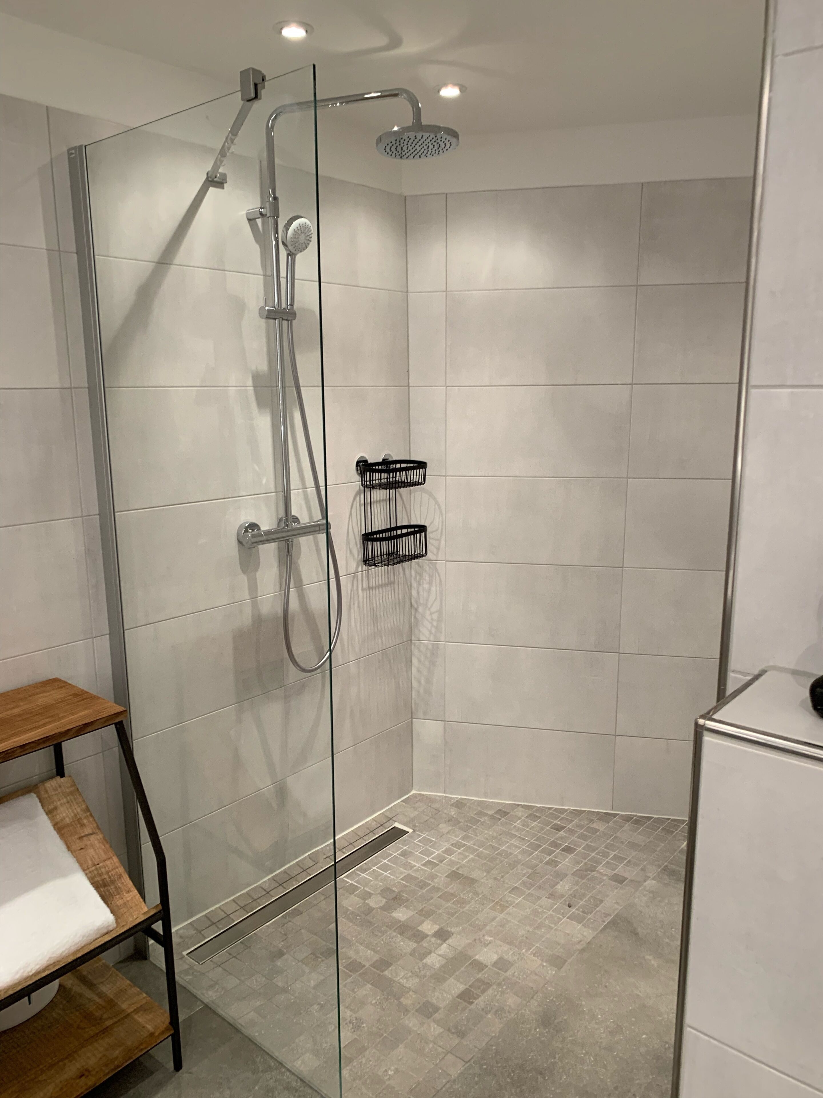Una ducha moderna con puerta de vidrio, cabezal de ducha manual y grifo empotrado en la pared. Se observa un pequeño estante de madera con una toalla blanca y una cesta negra.
