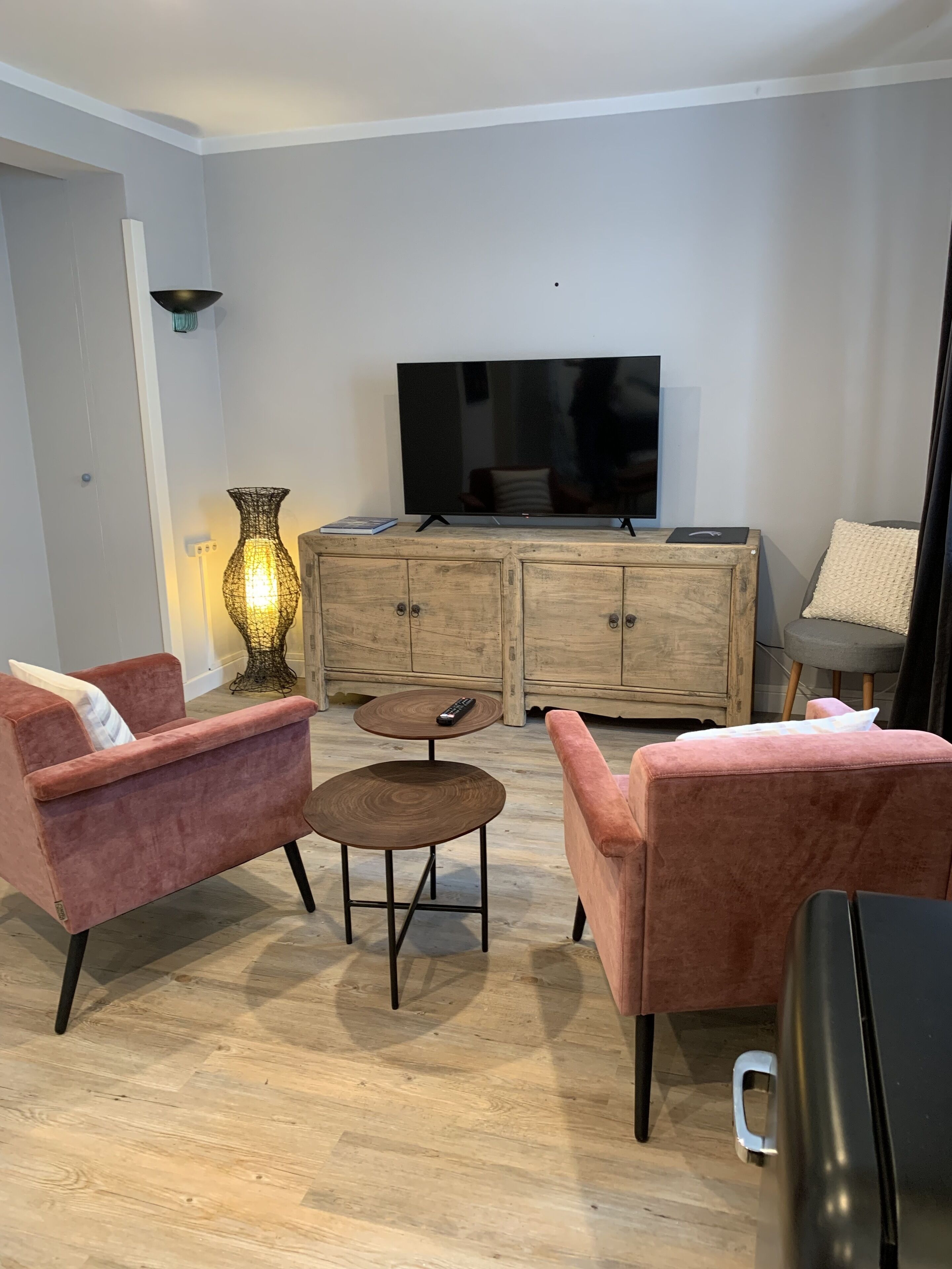 A living room with a flat-screen TV, two pink armchairs, a small round table, and a wooden cabinet.