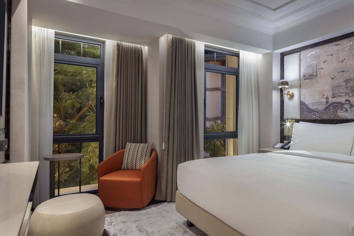 A hotel room with a large bed, a chair, a desk, and a view of greenery through the windows.