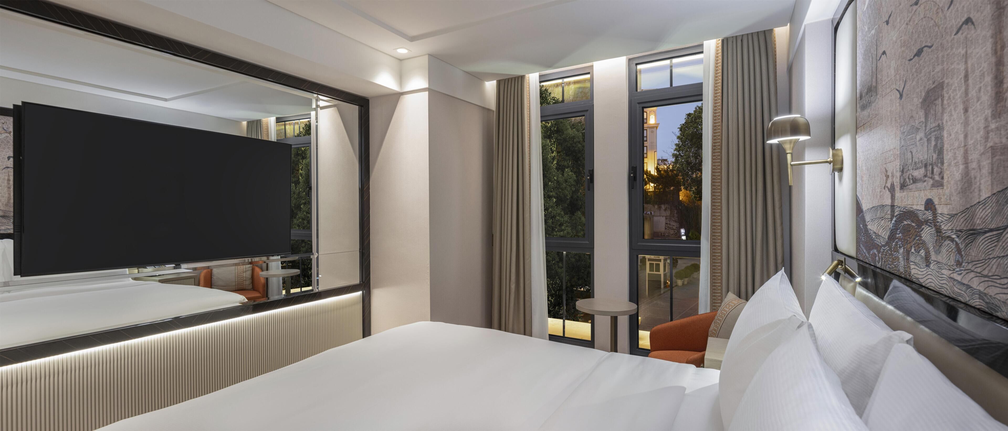 A hotel room with a large bed, a television, a window with curtains, and a view of the outside.