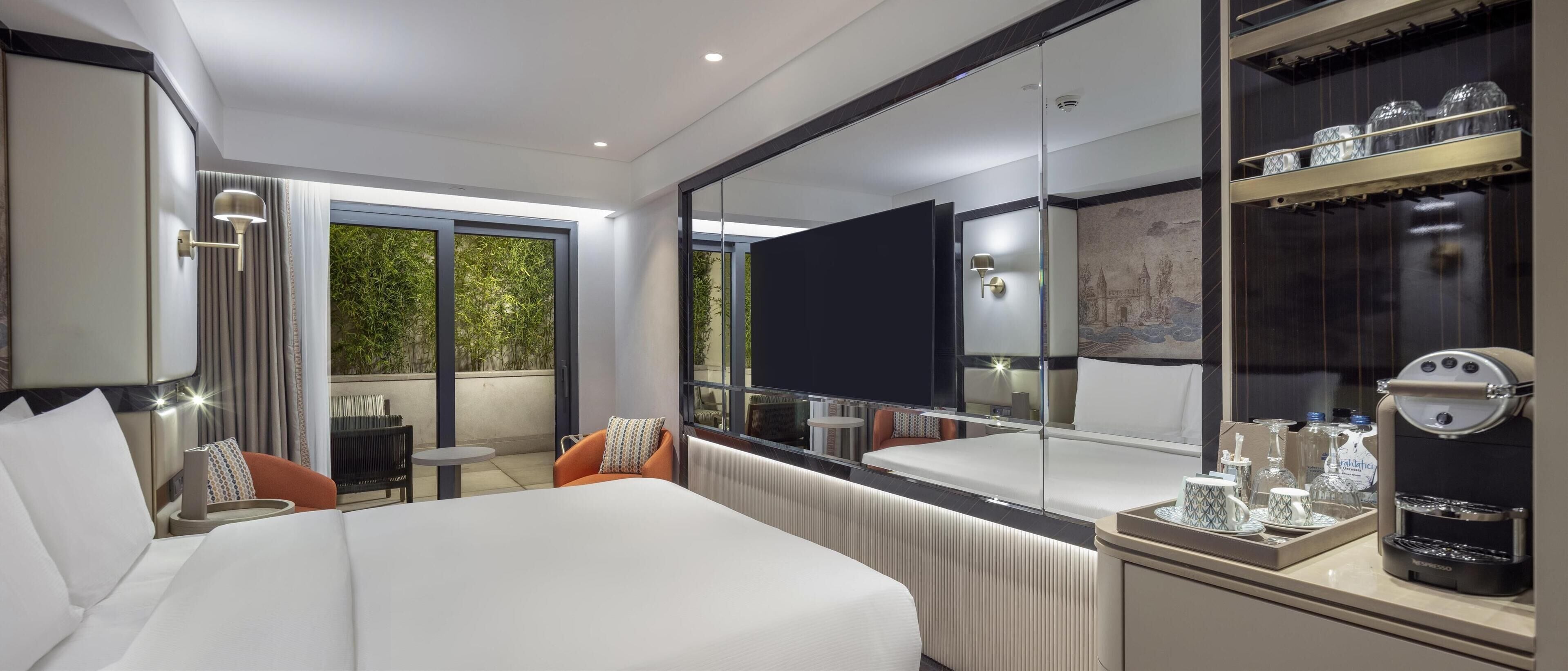 A modern hotel room with a large bed, a seating area with a table, a minibar, and a large mirror reflecting the room.