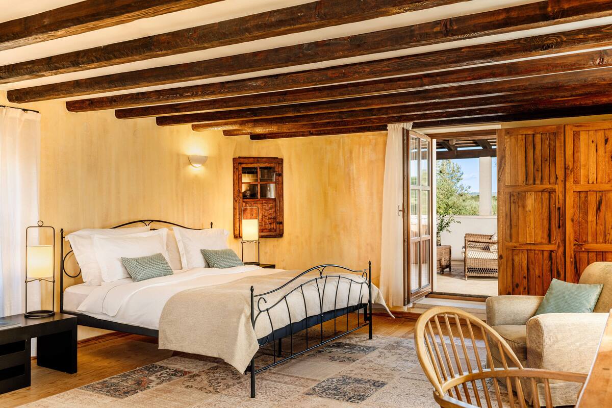 A bedroom with a bed, bedside table, lamp, and a view of the outside.