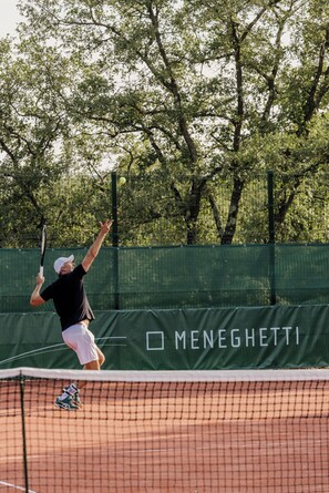 Een tennisser in volle beweging op een gravelbaan, met een groene omheining en bomen op de achtergrond.