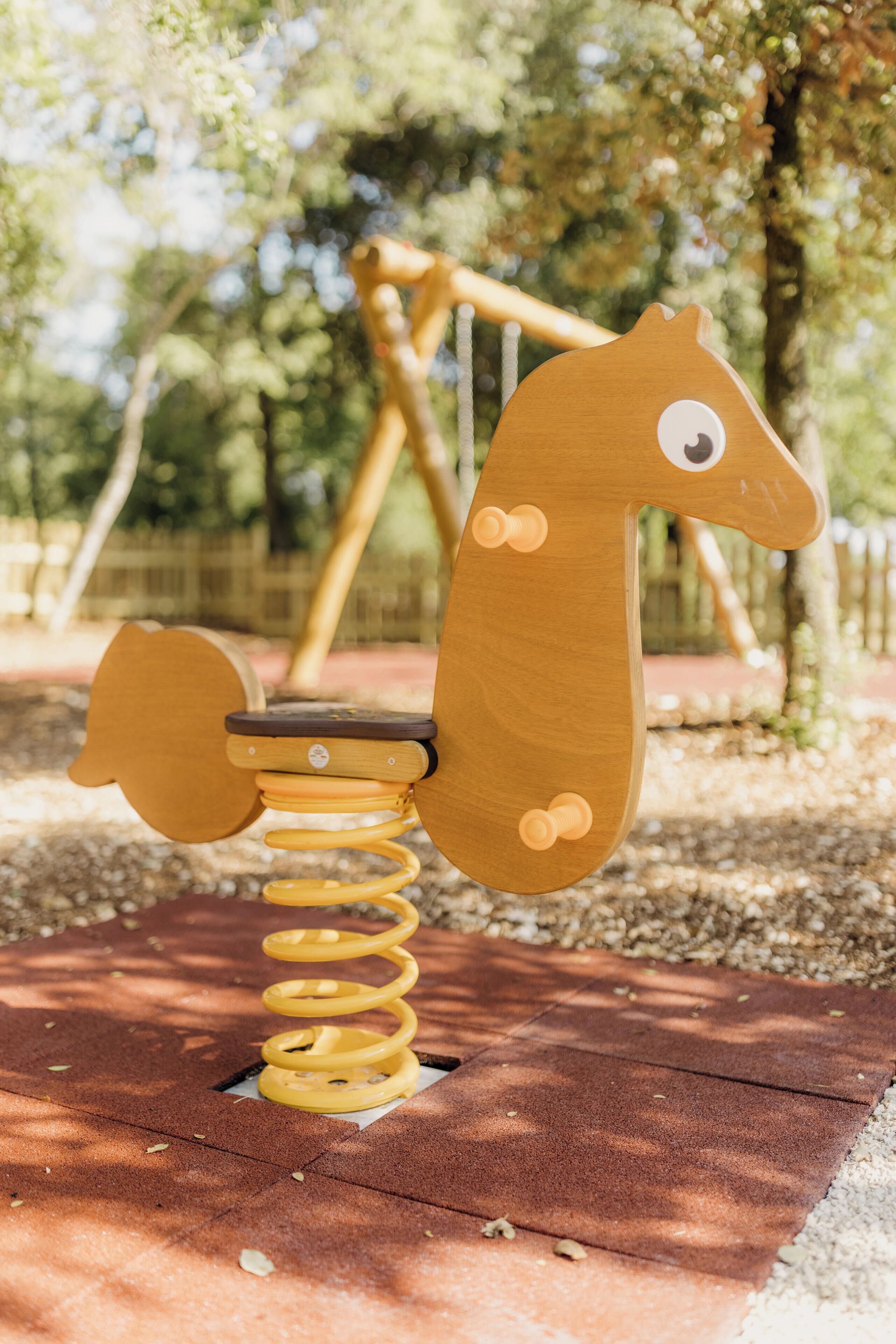 A wooden seesaw with a spring mechanism, set on a paved surface in an outdoor area with trees and a fence in the background.