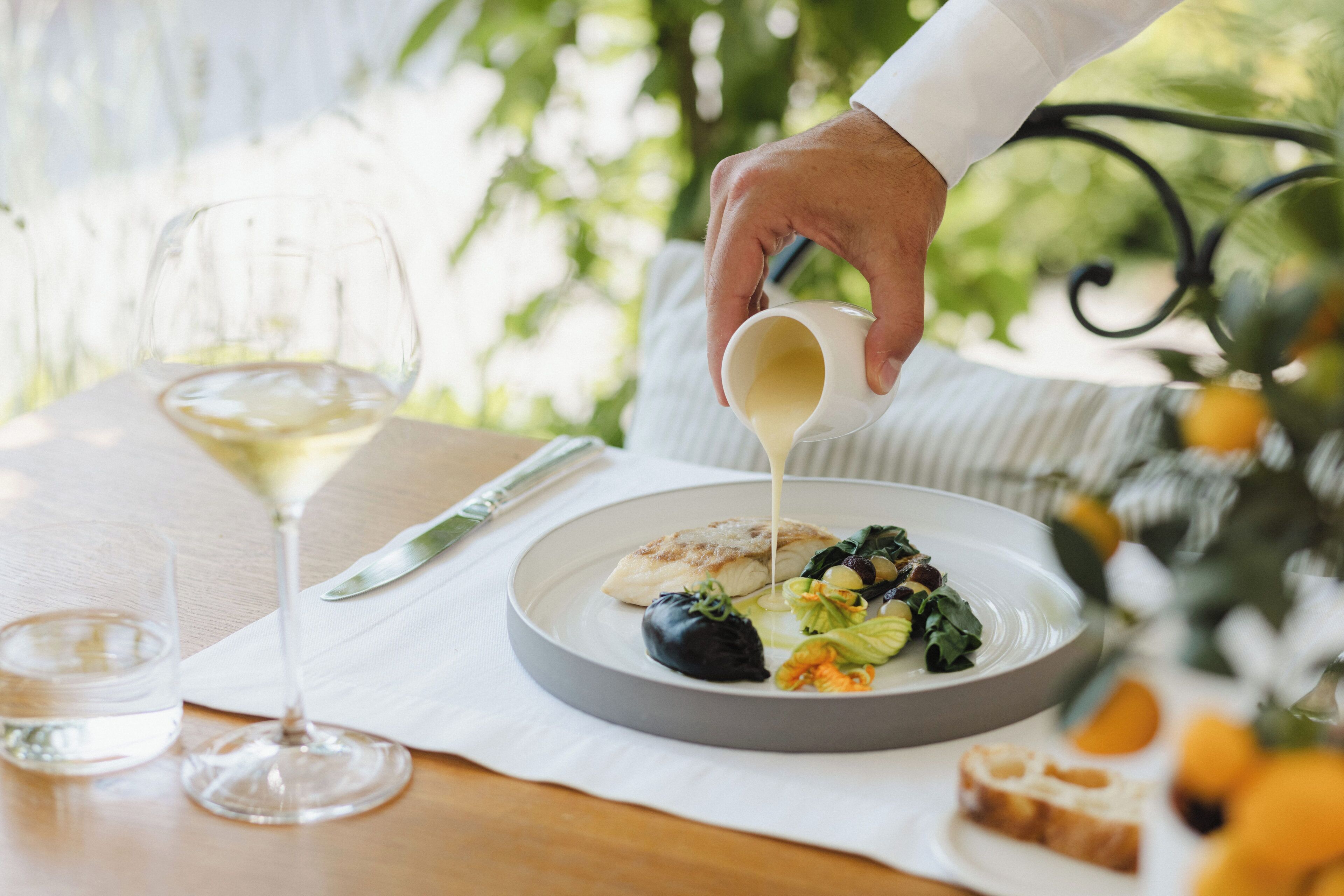 Une table à manger avec une assiette de nourriture, un verre de vin blanc et une main versant de la sauce.