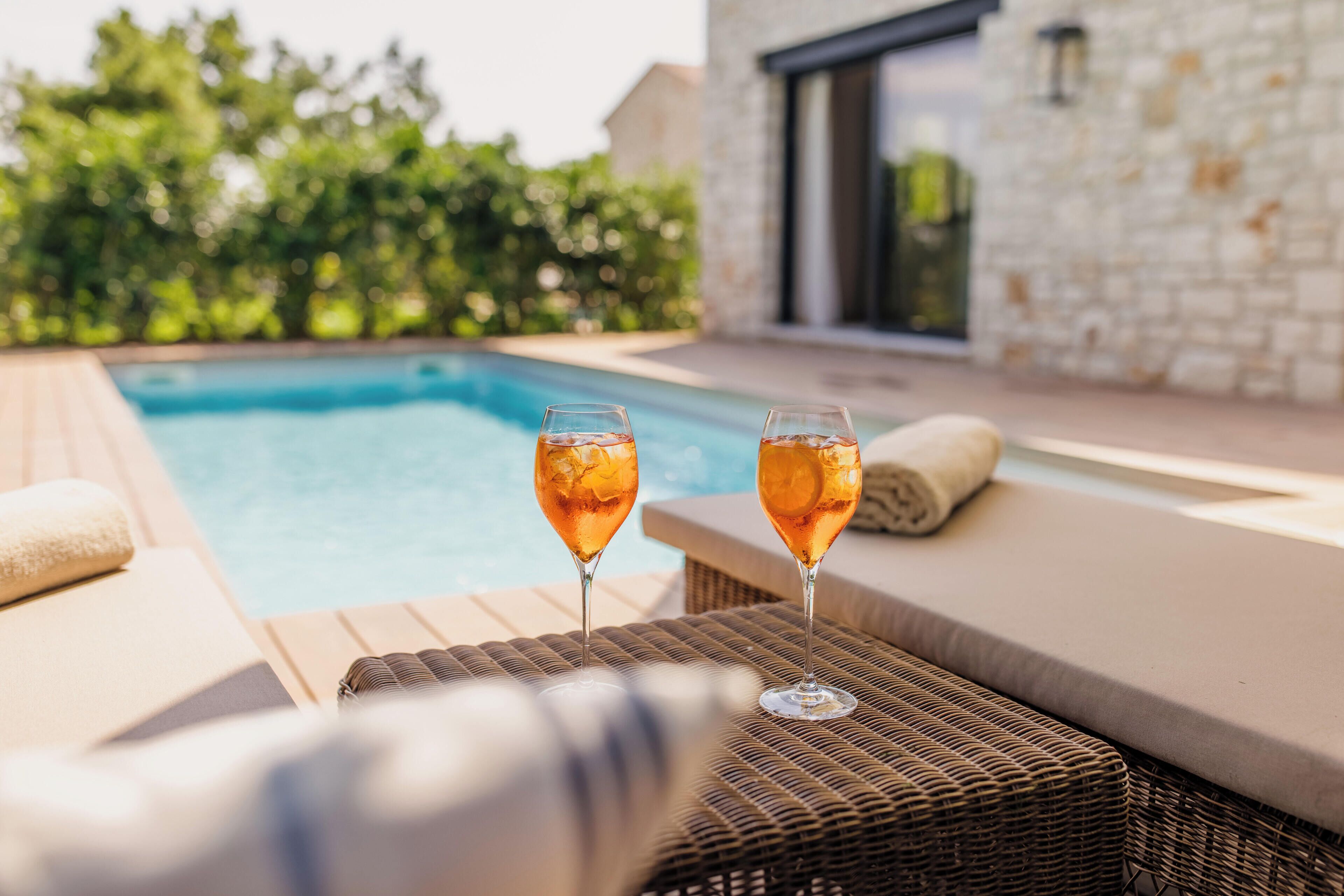 Escena junto a la piscina con dos vasos de una bebida, una silla de mimbre y un edificio de piedra al fondo.